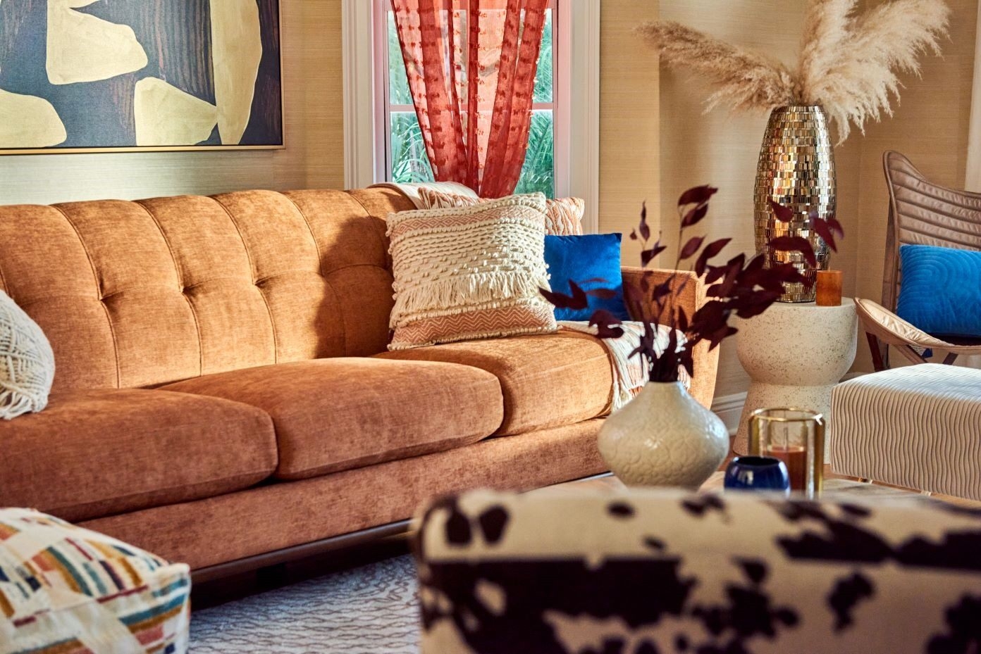 Orange sofa with decorative pillows, a cowhide ottoman, and a tall vase with pampas grass.
