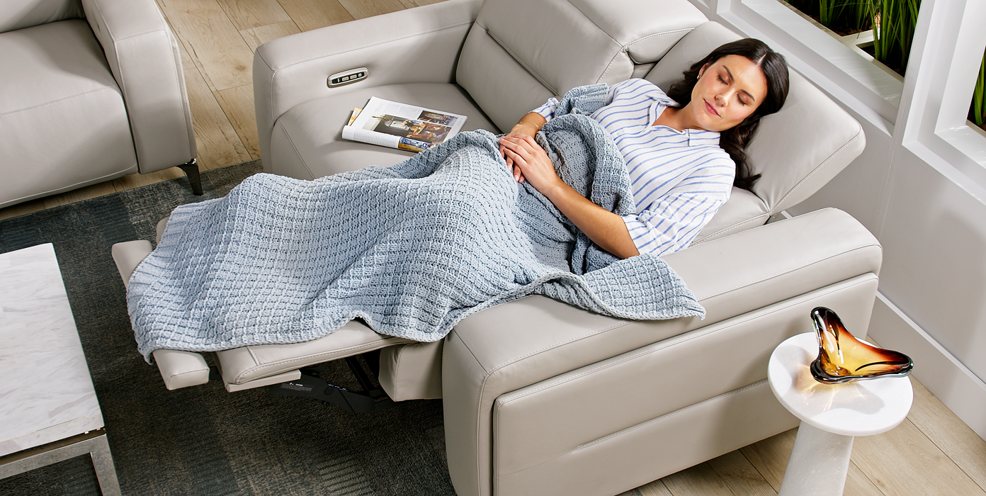 A woman sleeping on a light gray recliner sofa, covered with a blue blanket.