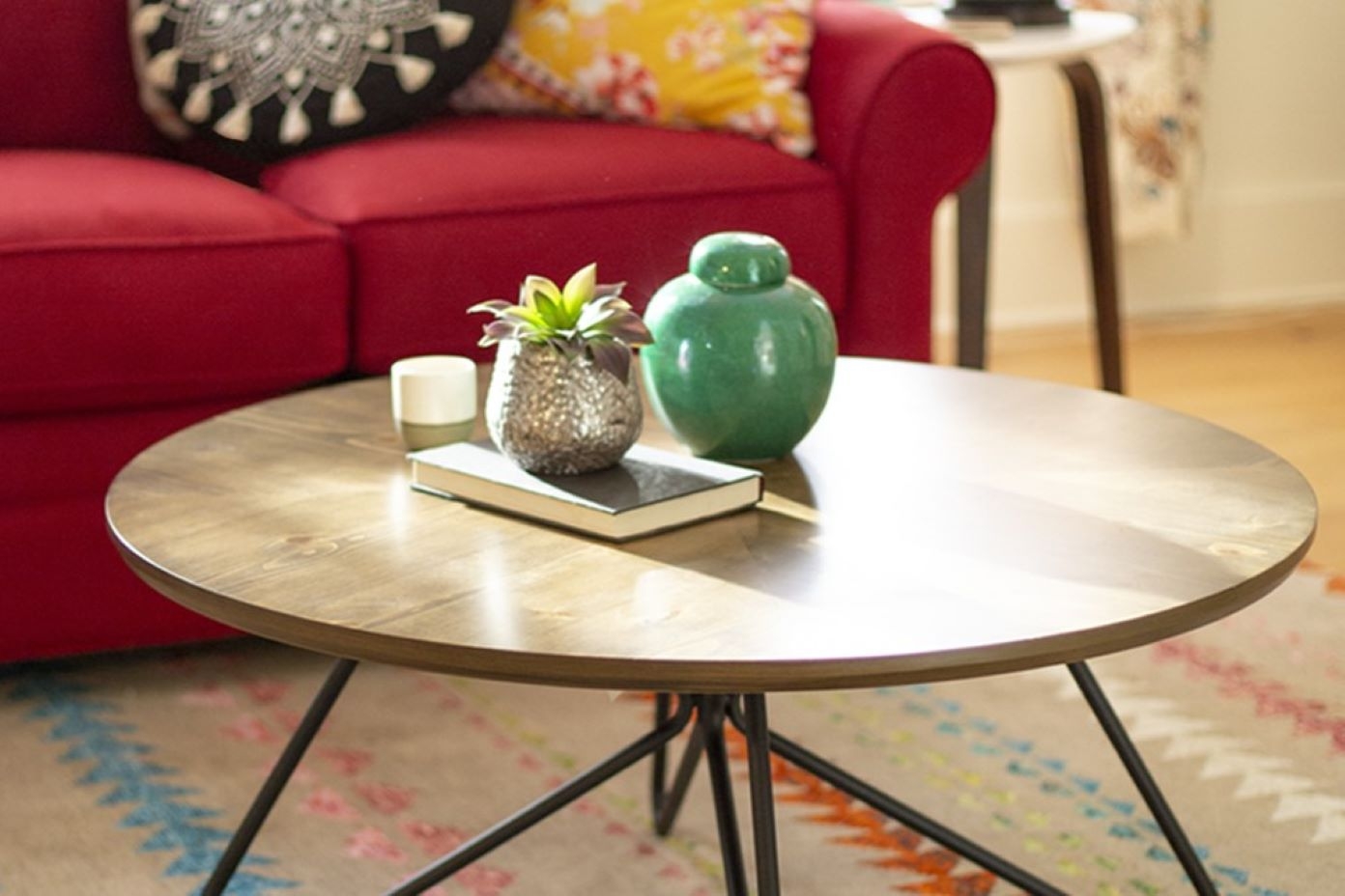 A round wooden coffee table with black legs, a red couch, and decorative items.
