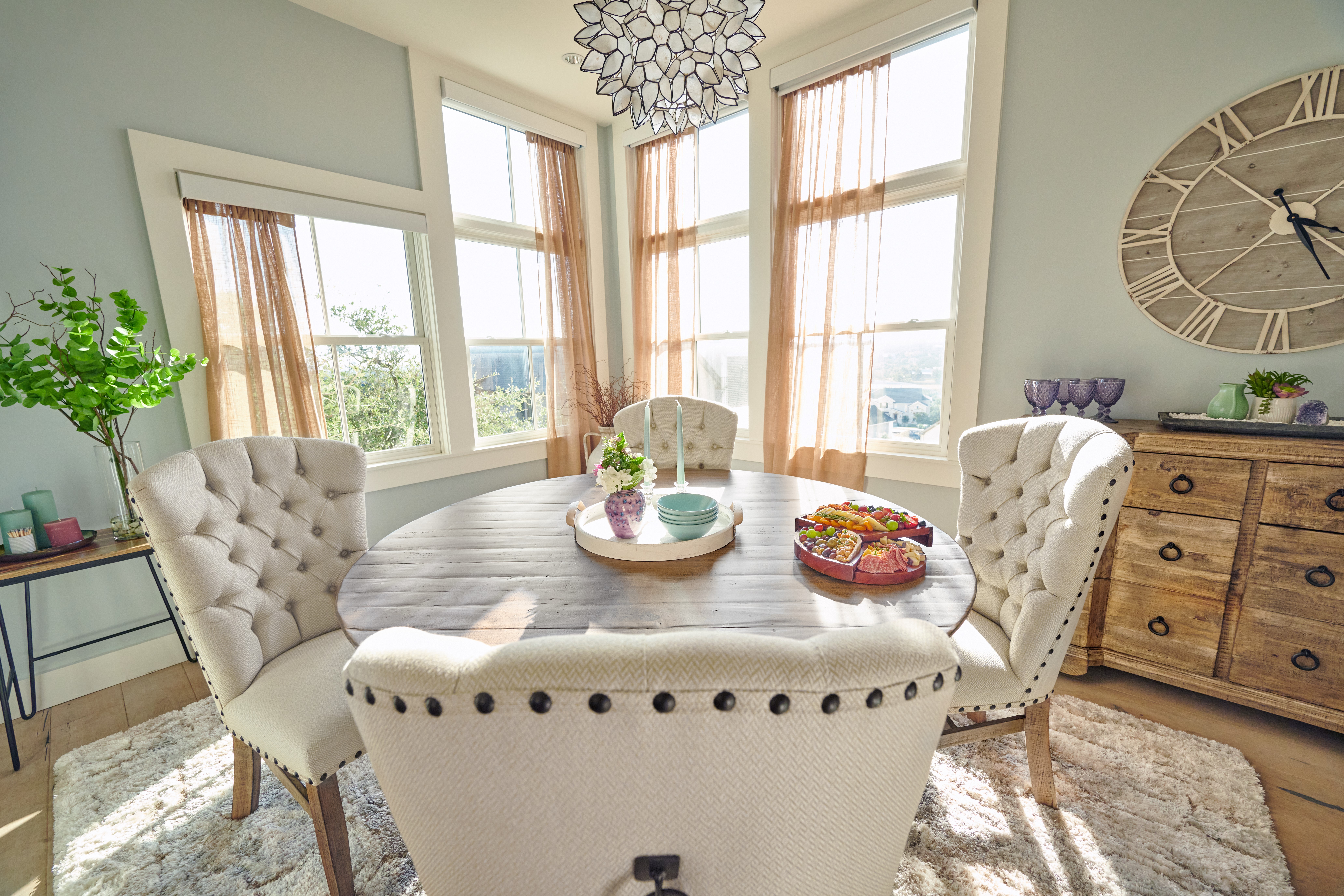 A dining room with a round wooden table, tufted chairs, and a large clock on the wall.