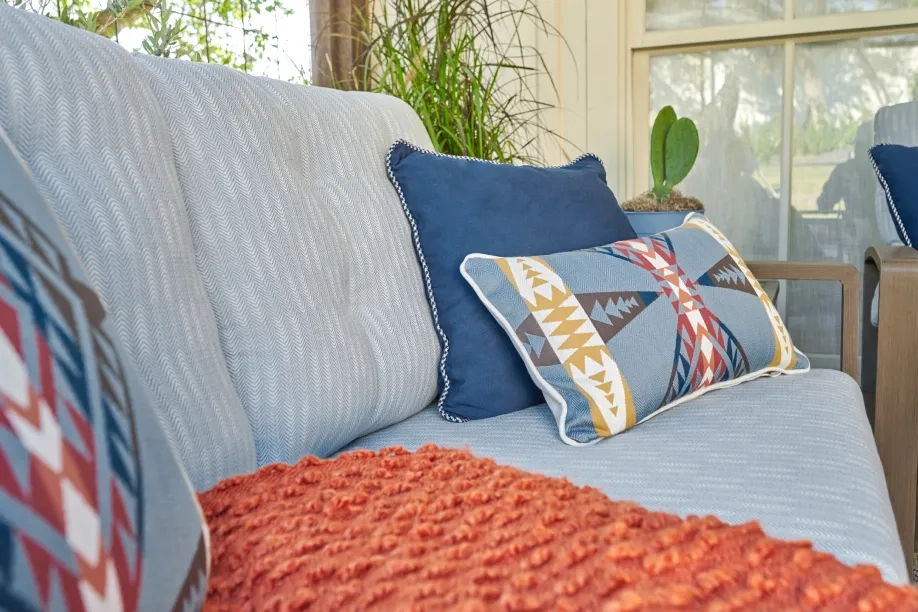 Outdoor sofa with blue and patterned pillows, orange knit blanket, and plants in the background