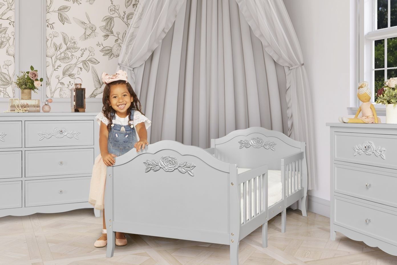 A young girl smiles while standing next to a gray toddler bed.
