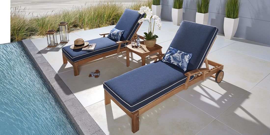 Photo of two teak chaise lounges with blue cushons next to a pool.