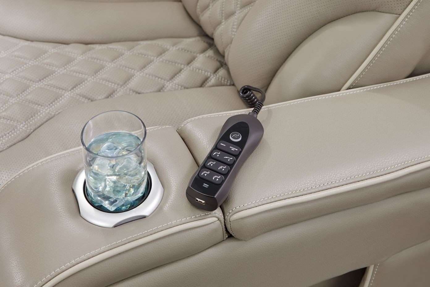 A close-up of a beige leather recliner with a cup holder, a glass of ice water, and a remote control.
