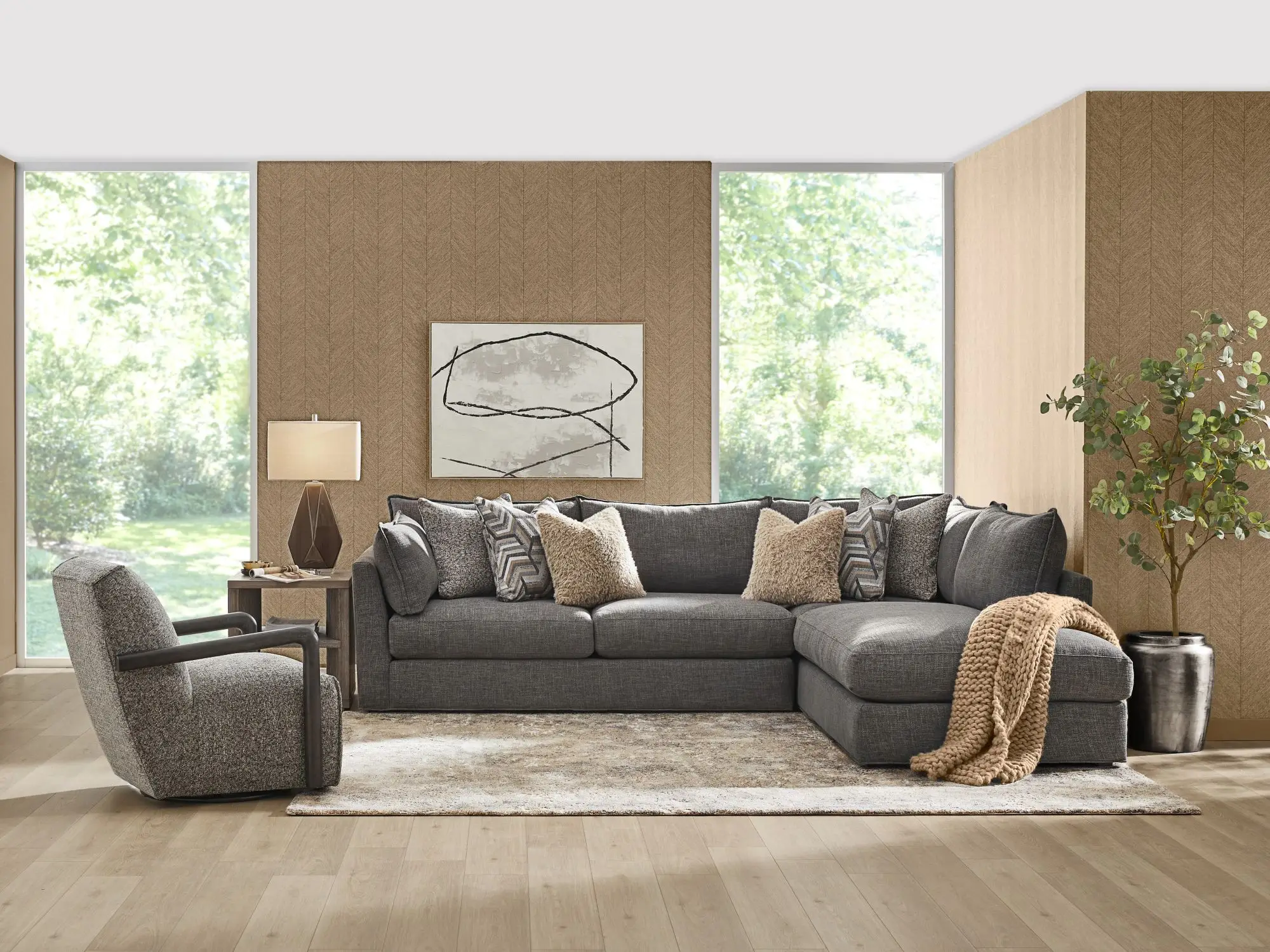 A gray sectional sofa with patterned and textured throw pillows, a matching armchair, and a potted plant in a living room.