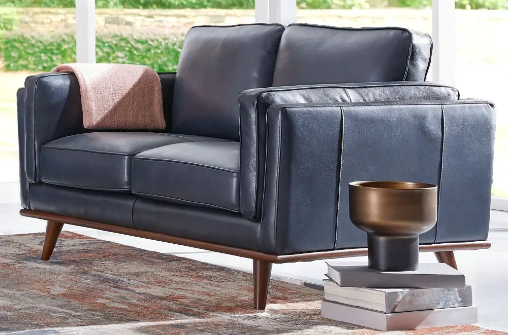 A dark blue leather loveseat with a mid-century modern wooden base, featuring a pink throw blanket and a decorative vase.