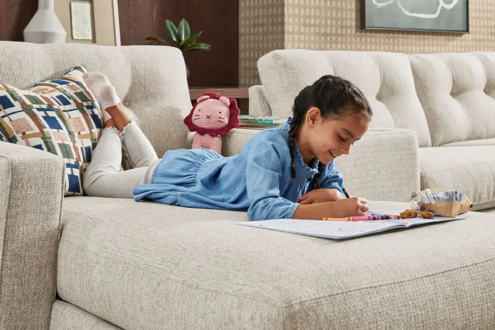 A young girl in a blue dress lies on a light-colored sectional sofa, coloring in a book next to a small plush lion toy.