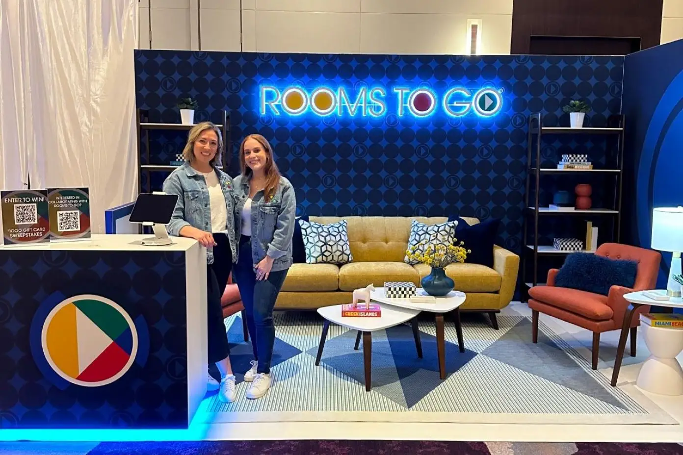 Two women stand in front of a Rooms To Go display. The display includes a sofa, chairs, and tables.
