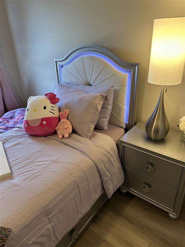 A bed with a silver headboard, a nightstand, and a lamp. There are pillows and stuffed animals on the bed.
