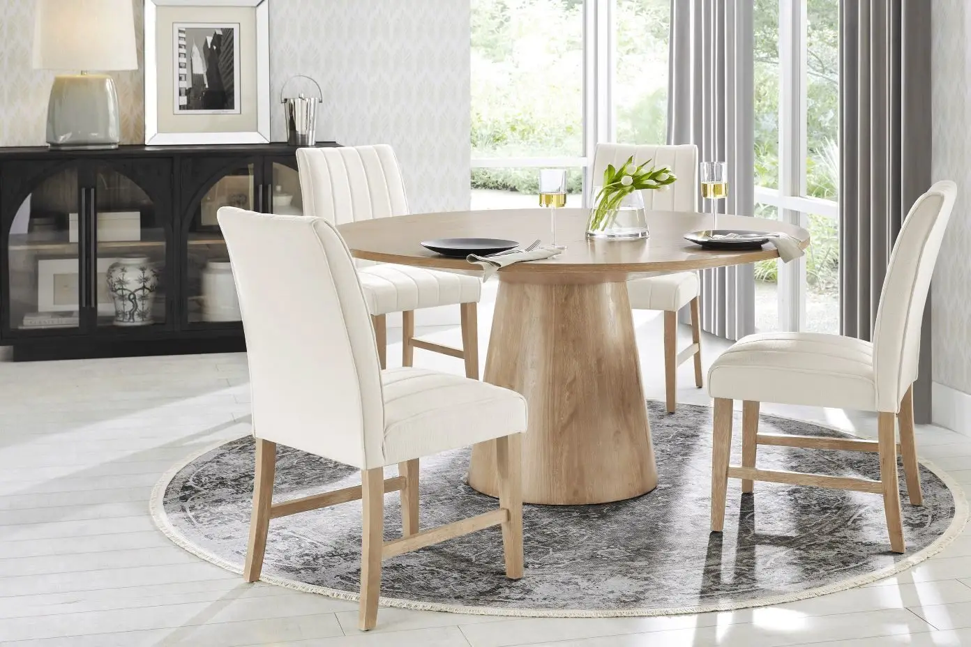 A dining room set with a round wooden table, four white upholstered chairs, and a patterned rug.