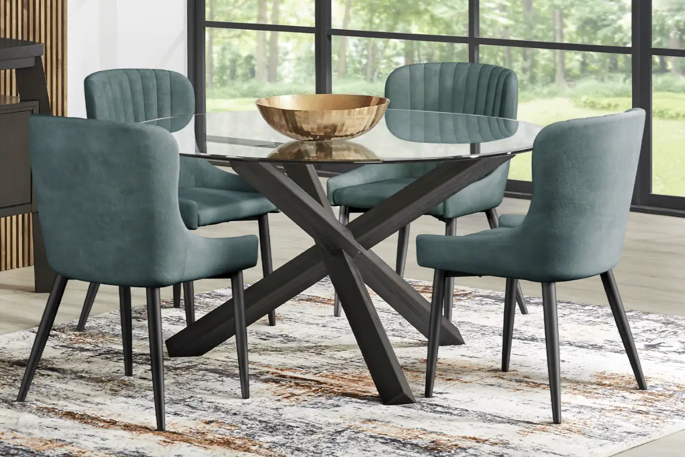 A round glass dining table with four teal chairs and a gold bowl on a patterned rug.

