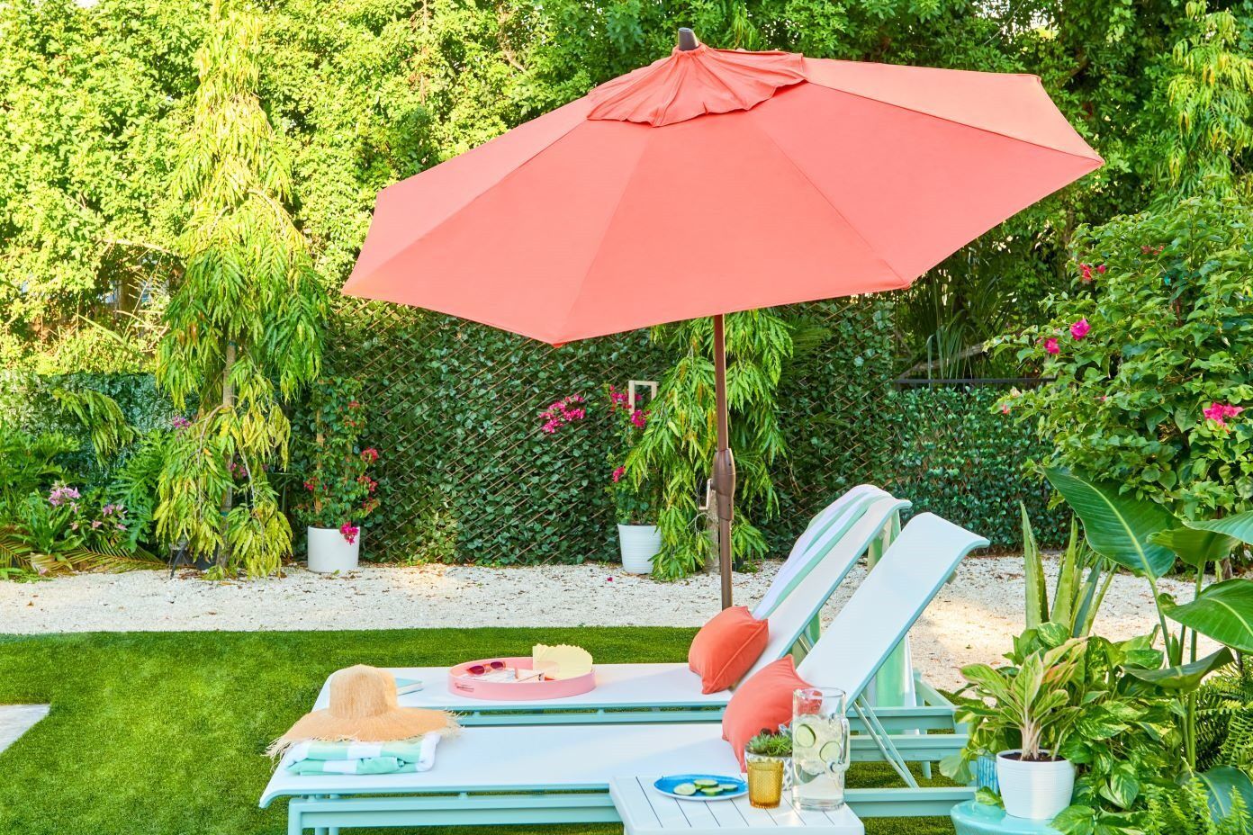 Two white lounge chairs with orange pillows under a coral umbrella in a garden.
