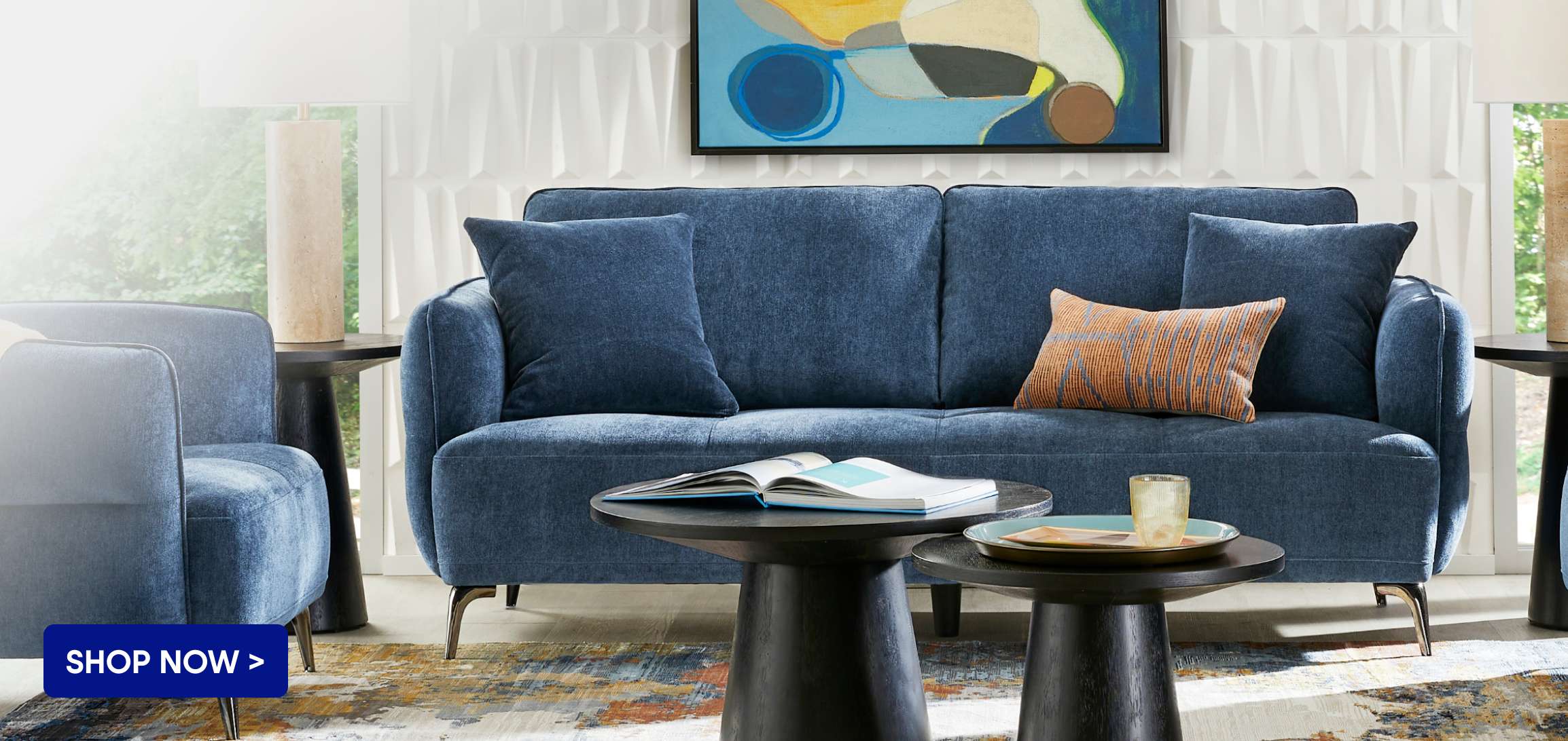 SHOP NOW > A blue sofa with pillows, two small black tables, and a painting.
