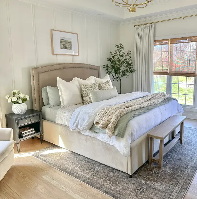 A cozy bedroom featuring a beige upholstered bed, sage green bedding, a chunky knit throw, and a wooden bench on a rug.