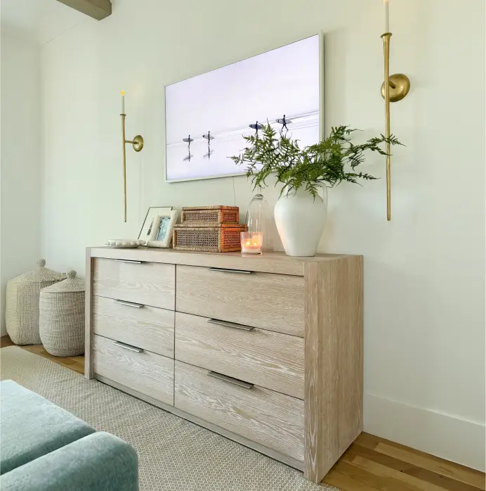 A light wood dresser with six drawers, topped with a vase of greenery, decorative boxes, and a wall-mounted television.
