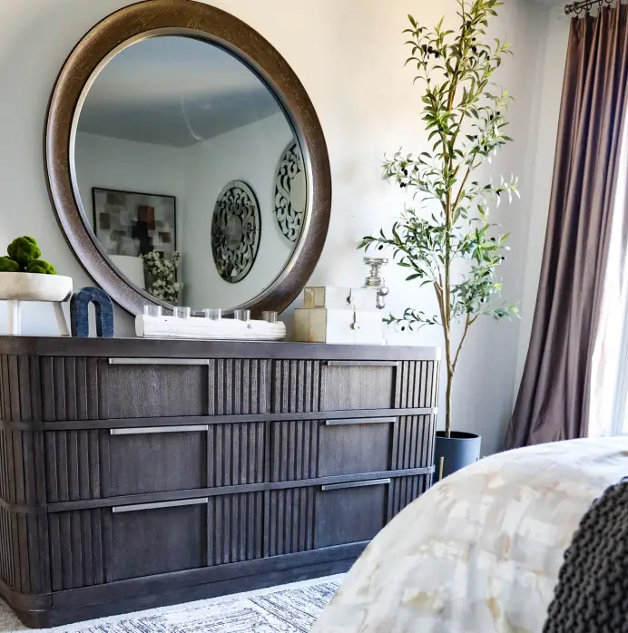 A dark wood, six-drawer dresser with vertical fluting, topped with a large round mirror, decorative boxes, and a tall plant.