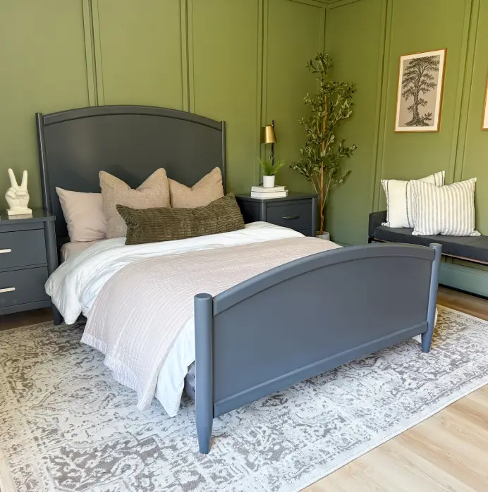 A dark gray bed frame with a light pink quilt and olive green accent pillow in a room with olive green paneled walls.