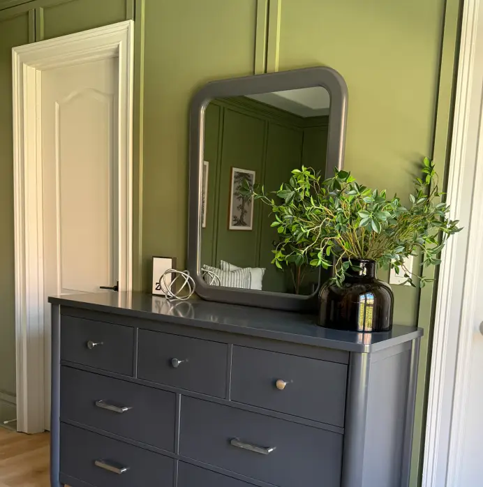 A dark gray dresser with a matching mirror sits against an olive green paneled wall, topped with a plant in a glass vase.