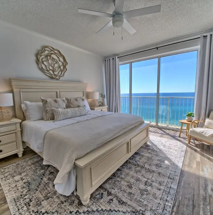 A bright, coastal-style bedroom featuring a large bed with neutral bedding, a patterned rug, and a balcony with ocean views.