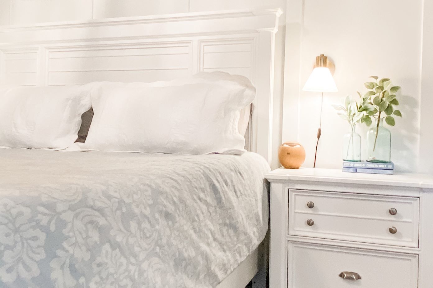 White bedroom with a bed, nightstand, lamp, and decorative vases.
