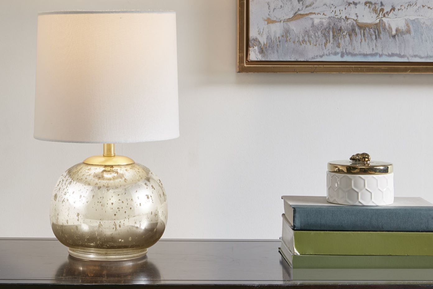 A table lamp with a gold base and a white lampshade, next to a stack of books and a decorative jar.
