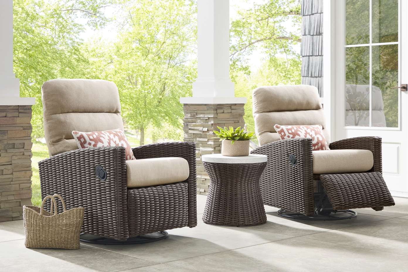 Two brown wicker recliner chairs with beige cushions and patterned pillows on a porch. A small side table and a basket are also present.
