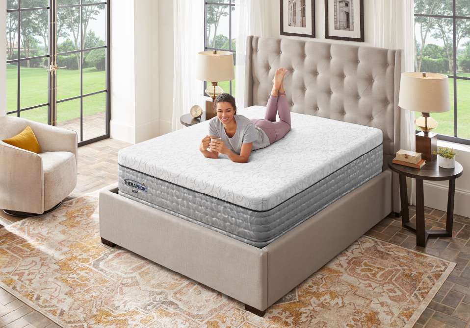 A woman is lying on a mattress, holding a mug. The mattress is on a bed frame.