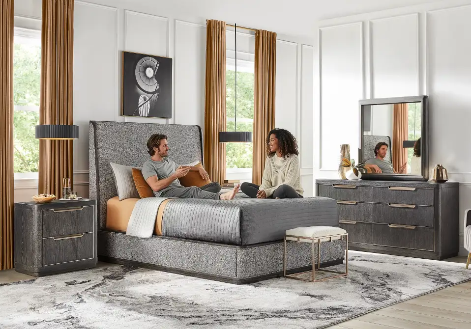 A bedroom with a bed, dresser, and two people sitting on the bed.