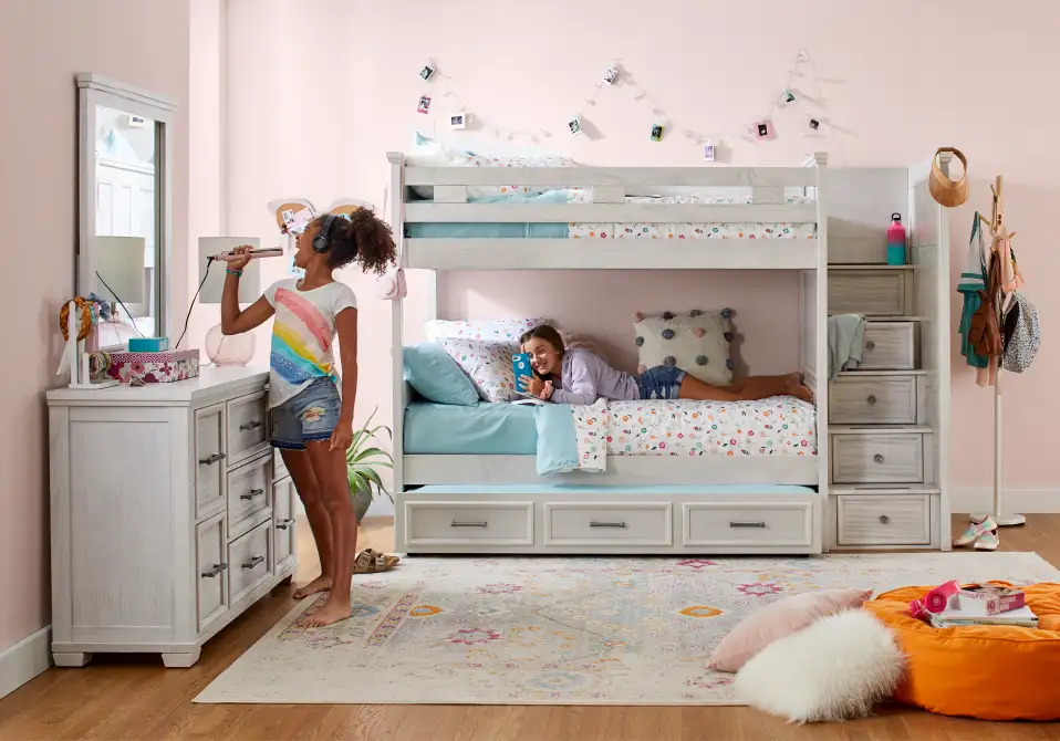 Two girls in a bedroom with a bunk bed, one singing into a hair straightener.