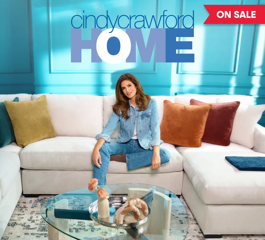 Cindy Crawford Home on sale. Cindy Crawford sits on a white sectional sofa in a living room setting.
