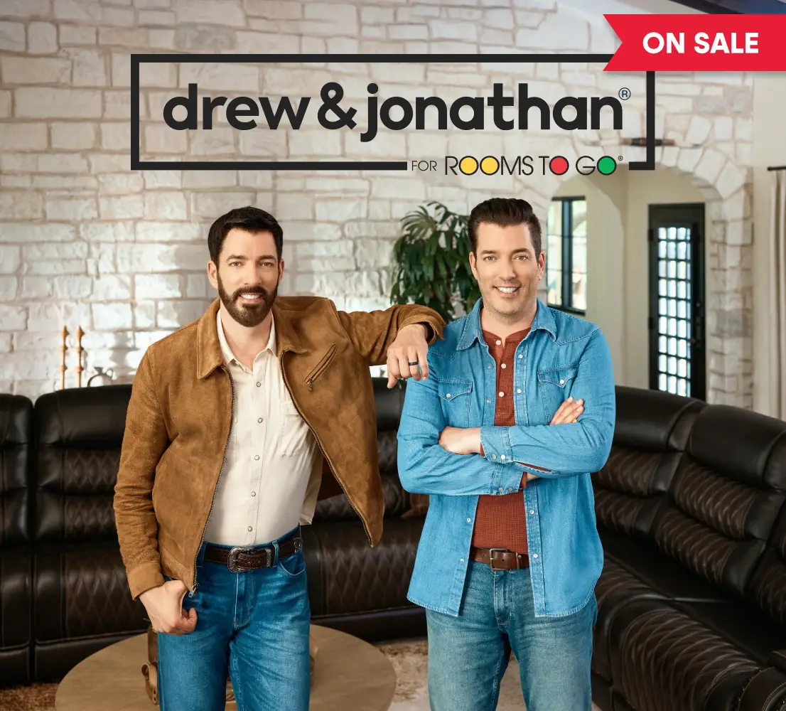 Drew & Jonathan for Rooms To Go, On Sale. Drew and Jonathan Scott stand in front of a dark brown leather sectional sofa.