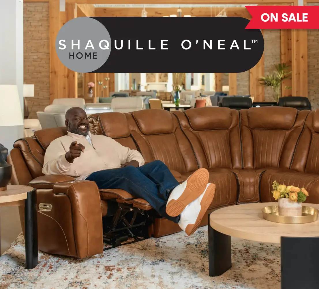 SHAQUILLE O'NEAL HOME ON SALE. Shaquille O'Neal sitting on a brown leather reclining sectional sofa in a furniture store.
