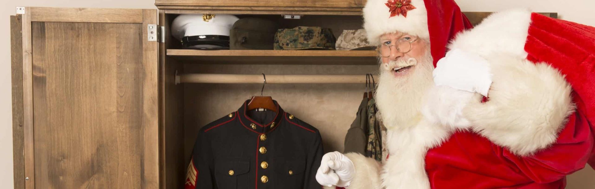 Santa Claus standing next to a wooden wardrobe with military hats and a uniform.