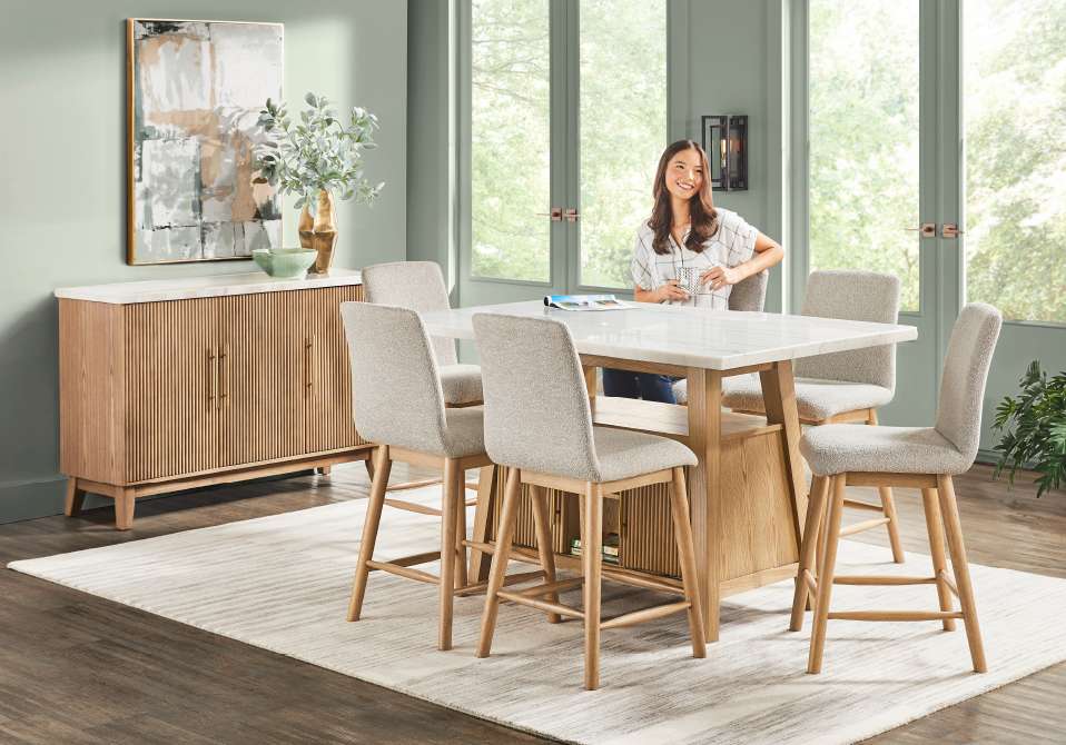 A dining room with a table, chairs, and a woman sitting at the table.
