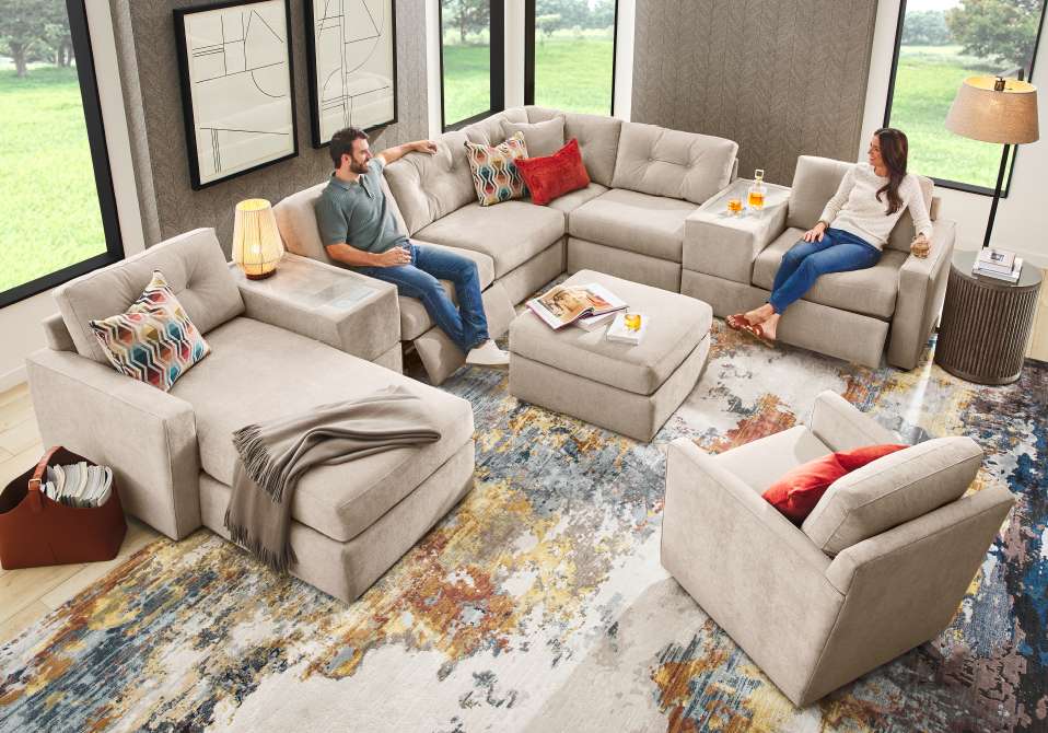 A beige sectional sofa with a chaise lounge, ottoman, and two armchairs on a colorful rug.

