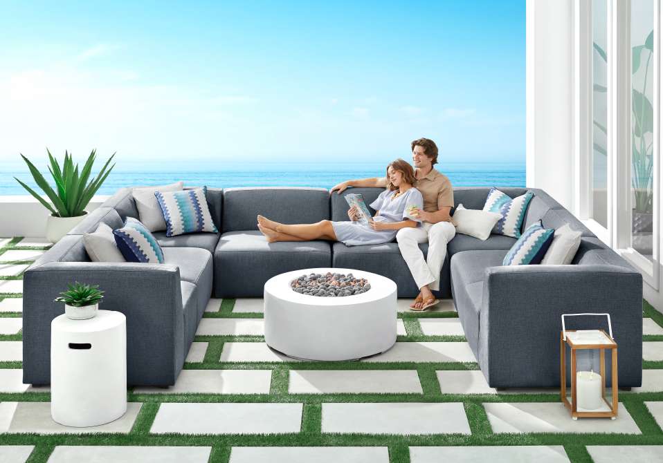 A couple relaxing on a gray sectional sofa on a patio overlooking the ocean.
