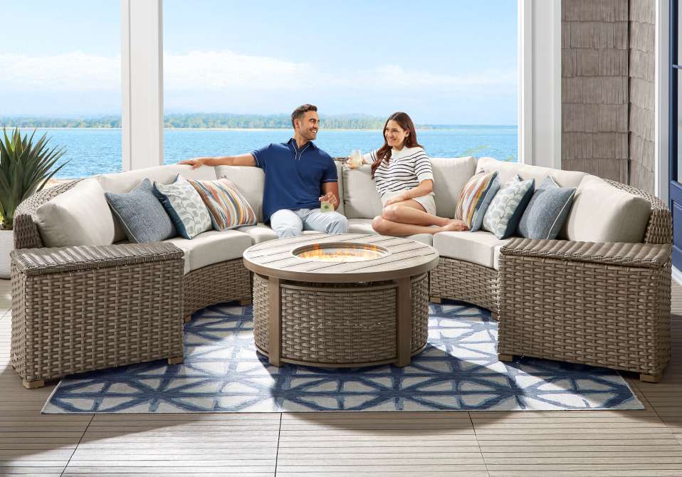 A couple sitting on outdoor sectional furniture with a fire pit and rug.
