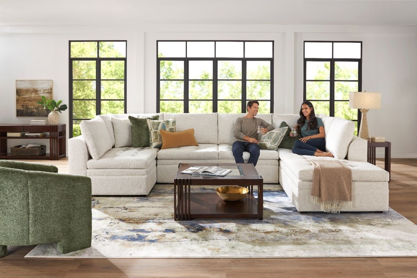 A living room with a white sectional sofa, a green armchair, and a couple sitting on the sofa.

