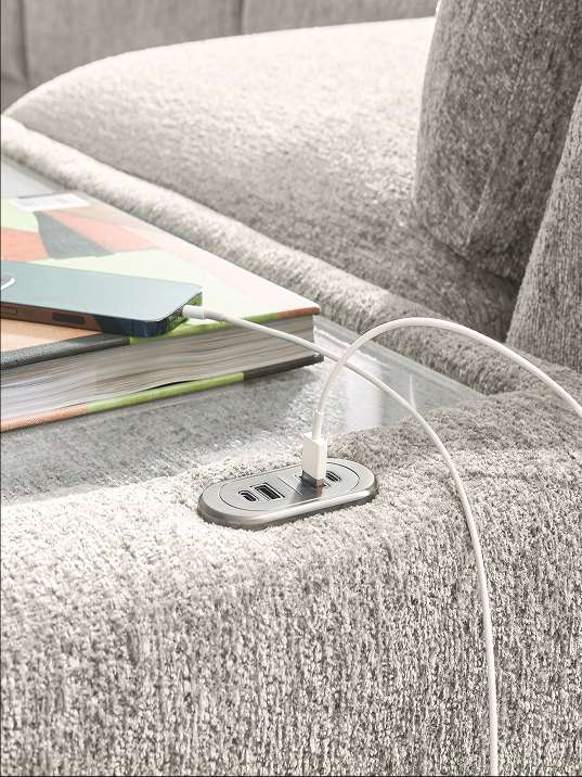 A close-up of a gray sofa with a built-in USB charging station. A phone is charging on a book.
