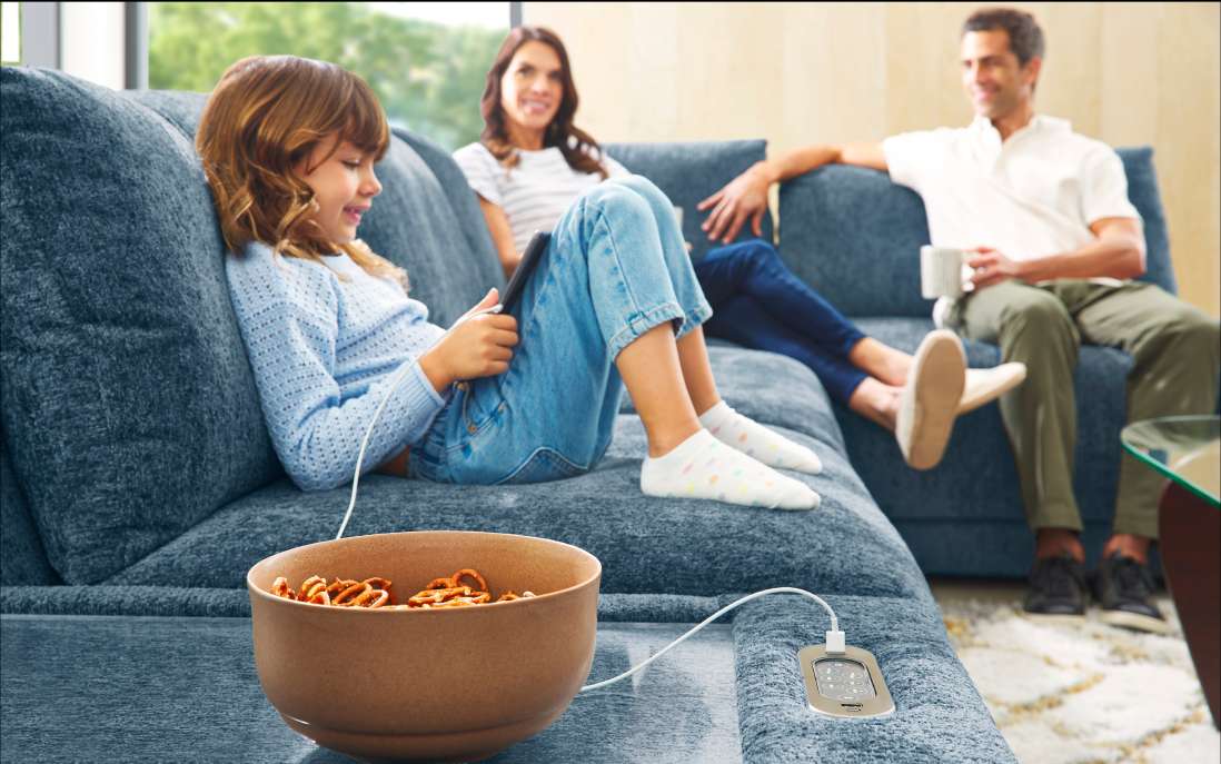 A family relaxing on a blue sofa. A girl is using a tablet, and a bowl of pretzels is nearby.
