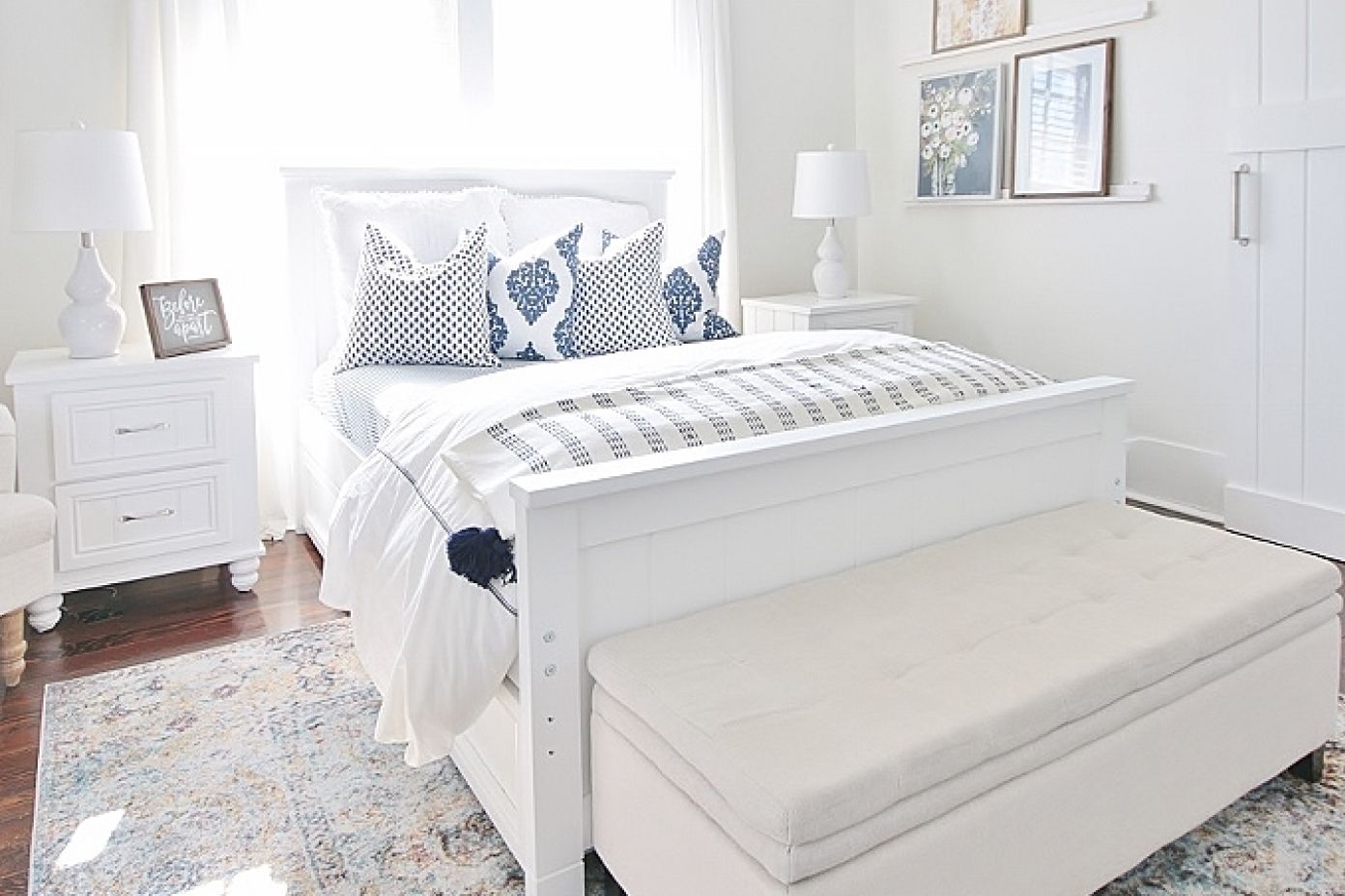 White bed with blue and white pillows, a white bench, and a patterned rug.

