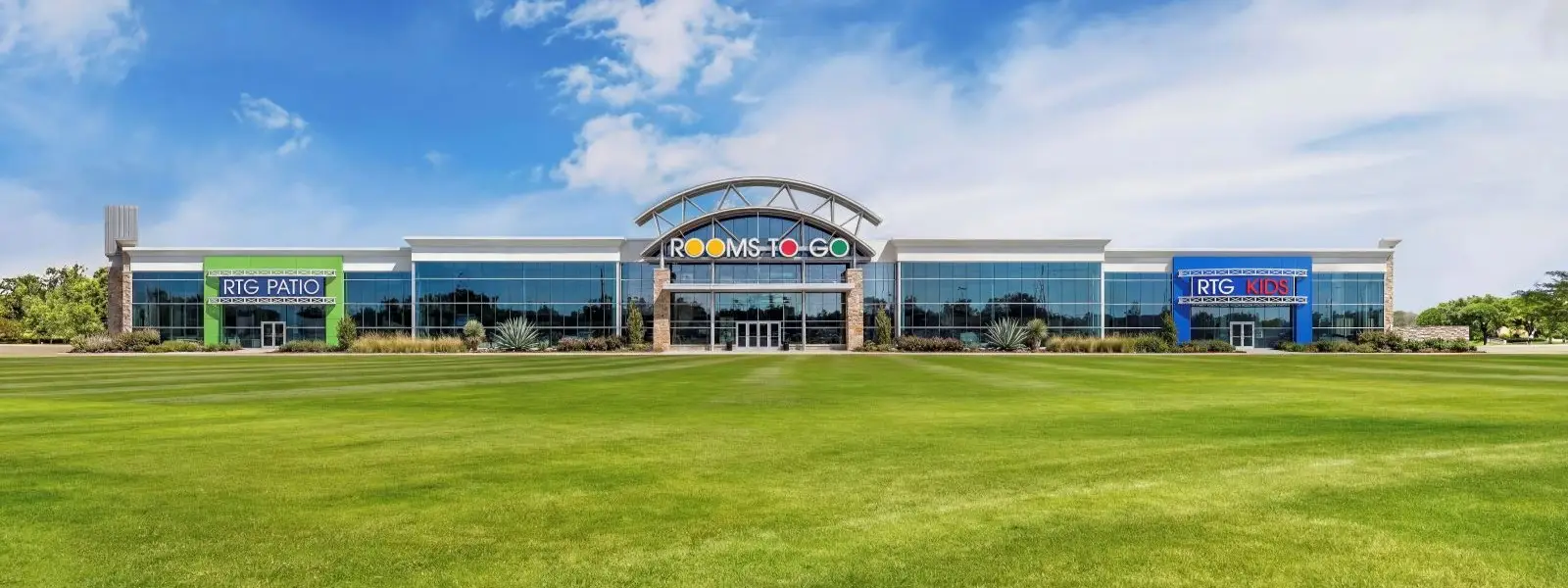 Rooms To Go, RTG Patio, and RTG Kids store exterior with a large green lawn in front under a blue sky.
