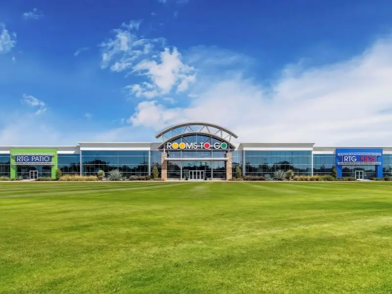 Rooms To Go store exterior with RTG Patio and RTG Kids sections, featuring a large green lawn under a blue sky.