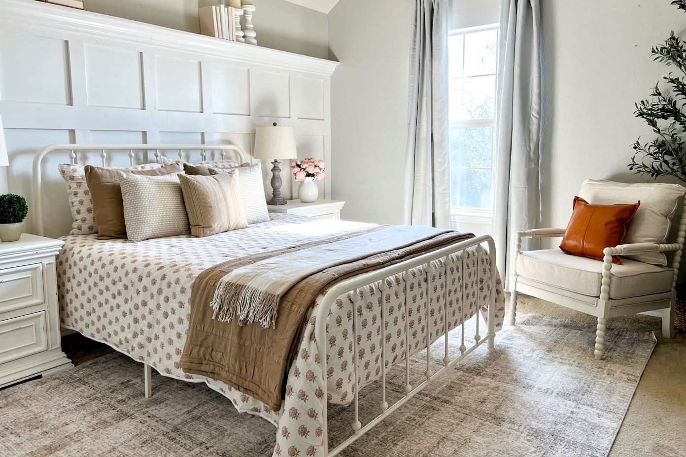 Bedroom with a white metal bed, patterned bedding, and a white armchair with an orange pillow.
