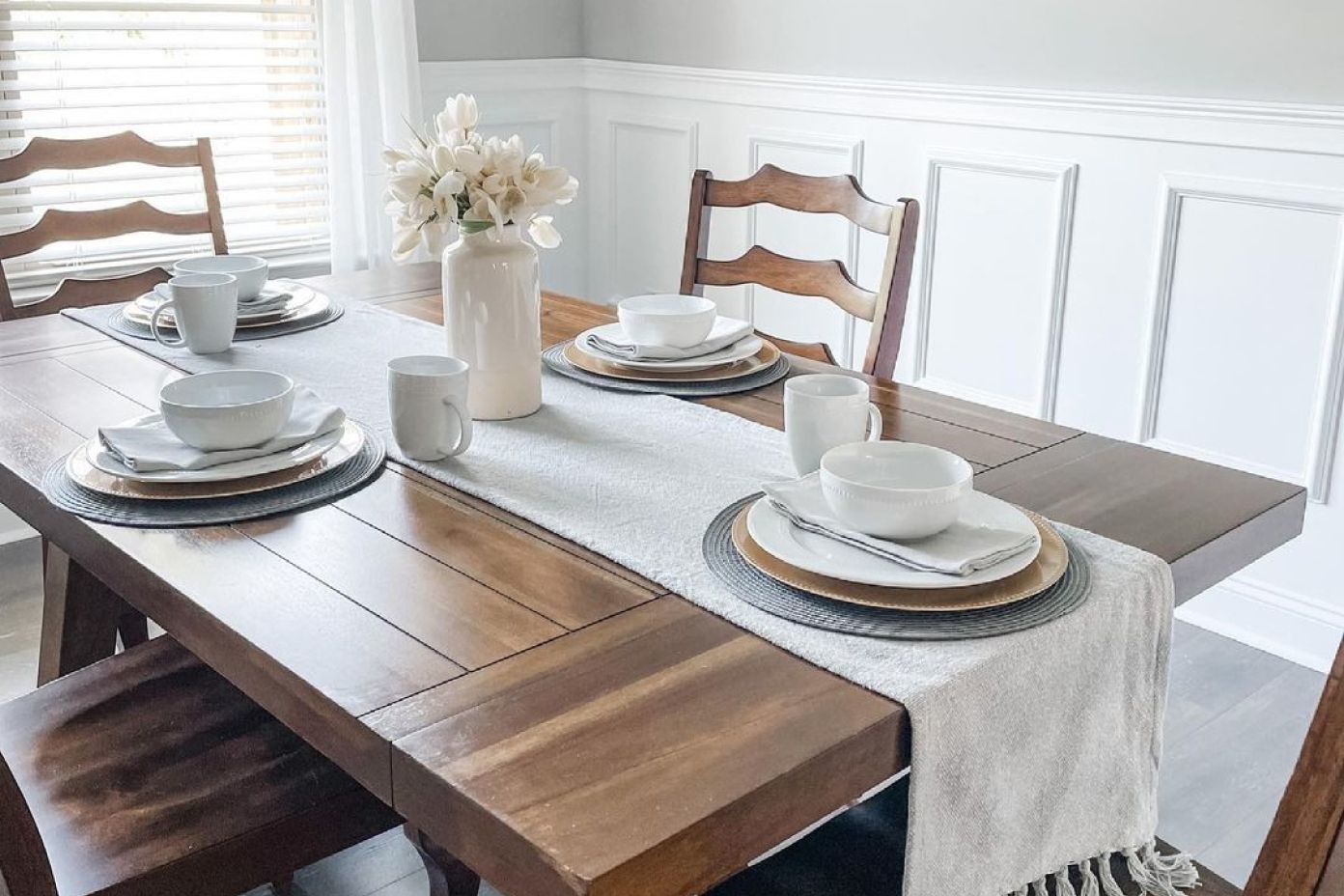 A wooden dining table set for a meal with white dishes and a vase of flowers.
