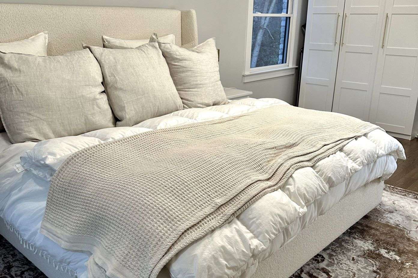 A bed with white bedding, beige pillows, and a beige waffle-knit blanket.
