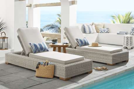 Two outdoor wicker chaise lounges with white cushions and blue striped pillows near a pool.
