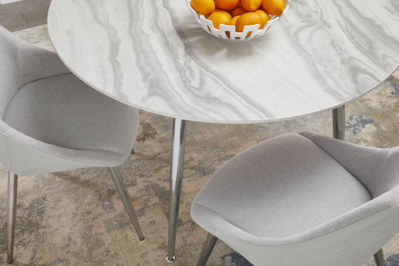 A round marble table with two gray chairs and a bowl of oranges.
