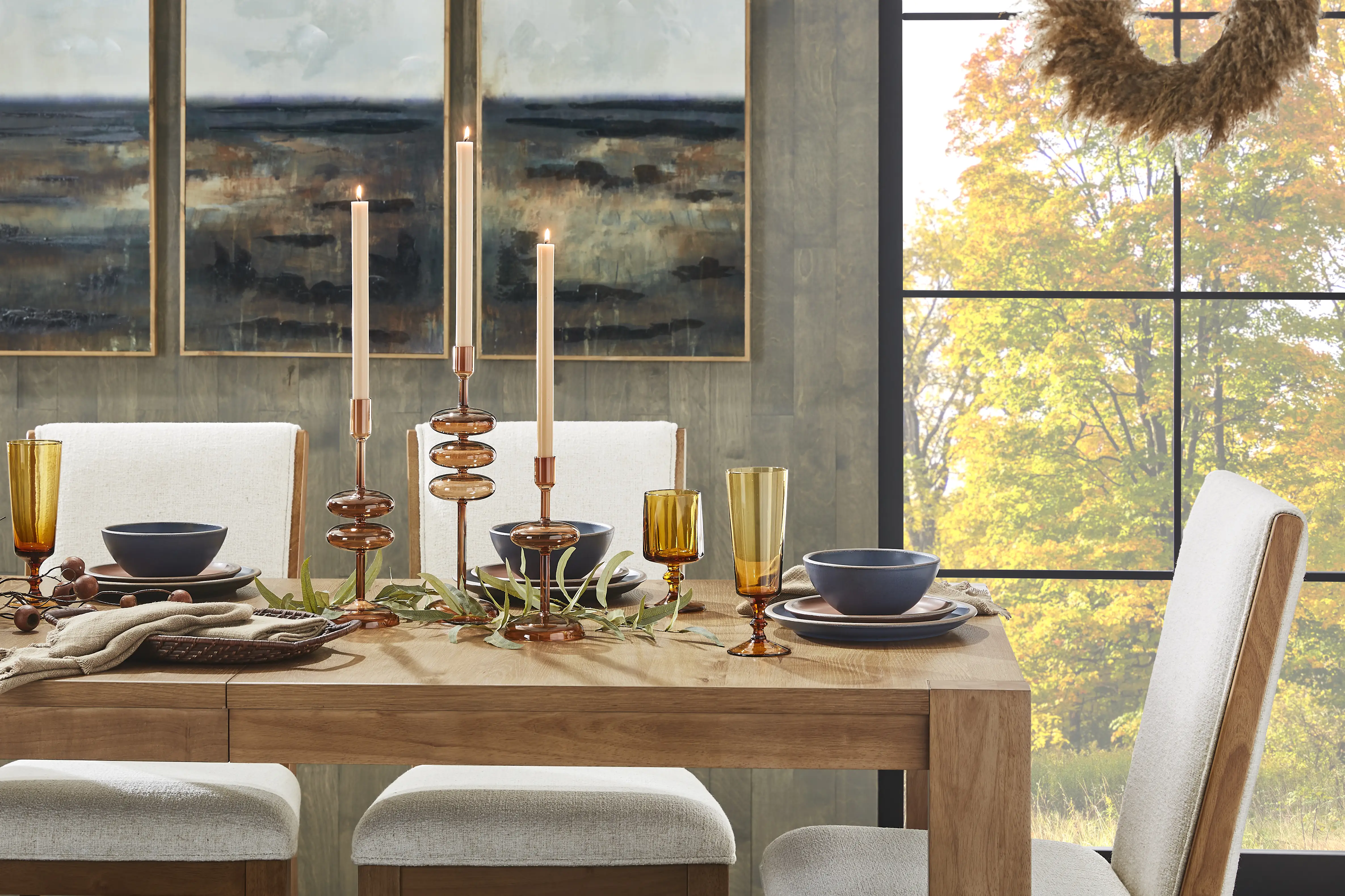A dining table set for a meal with candles, glassware, and a view of fall foliage.
