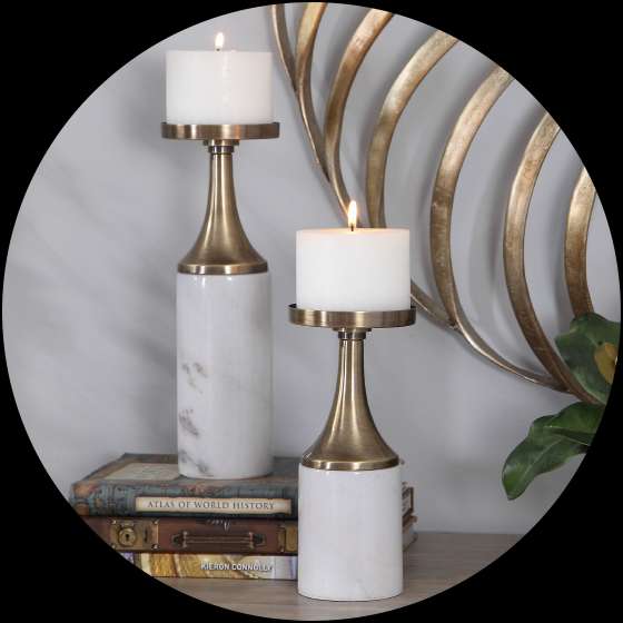 Two candle holders with lit candles on a wooden surface, with books and a gold wall decoration in the background.
